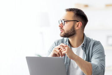 Bored young guy sitting near laptop, looking aside, daydreaming, thinking about something during online work at home office. Millennial Caucasian man having dull remote job