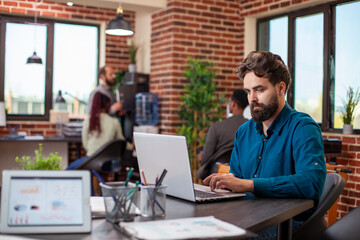 Fototapeta premium Caucasian businessman works on business strategies using his laptop in brick wall workspace. Bearded male analyst seated at the table researching and reviewing company project plans in startup office.