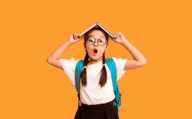 Funny Asian Elementary School Girl Posing With Book On Head Standing Over Yellow Background. Studio...