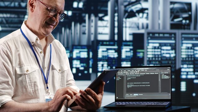 Elderly worker in data center, solving tasks on tablet while running scripts on laptop terminal. Certified engineer amidst servers, accessing databases and manipulating lines of code