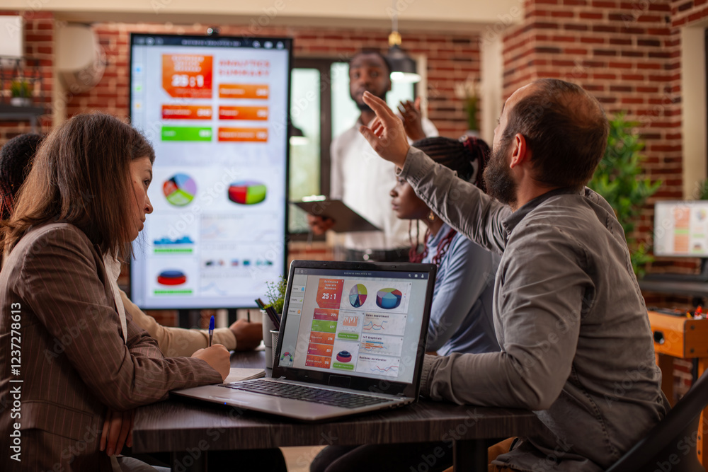 Poster white man raising his hand during meeting with laptop displaying business analytics nearby on table.