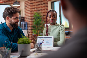 Black entrepreneur guiding her colleagues with market strategy insights, utilizing a digital device. Female startup leader using a tablet to explain company goals to her team in brick wall office.