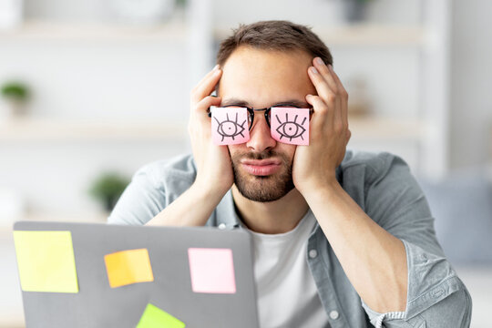 Tired young man having stickers with open eyes drawing on his glasses, sleeping in front of laptop at home office. Exhausted millennial man overworking, napping, failing to meet deadline