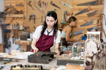 Interested young woman in pinafore examining metal template in carpentry workroom