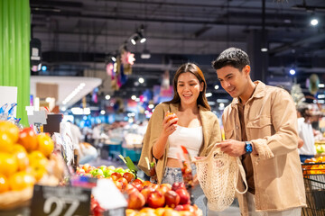 Happy Asian family couple using reusable cotton shopping bag buying food at supermarket. Man and woman enjoy sustainable living and healthy lifestyle choosing organic vegetable and fruit on shelves.
