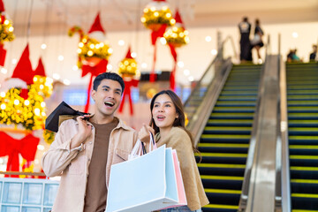 Happy Asian family man and woman shopping and buying goods at department store. Couple enjoy urban lifestyle holding shopping bag standing on escalator in shopping mall on summer holiday vacation.