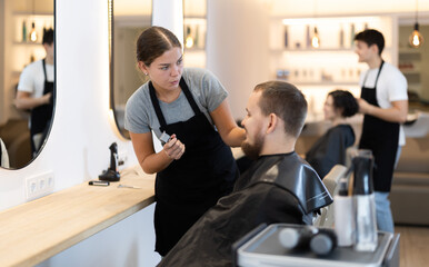 Young woman barbershop employee stands next to male client, discussing details of haircut. Stylist...