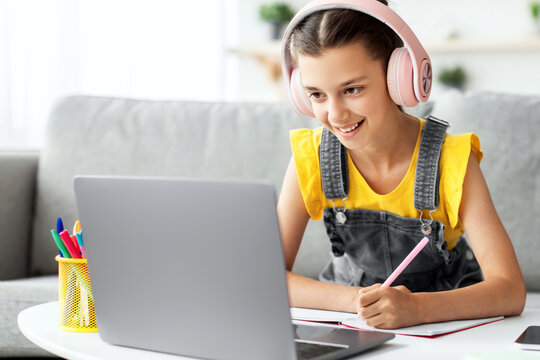 Educational Lecture. Portrait of smiling girl in pink headphones sitting on couch at table using laptop looking at screen, writing in her copybook taking notes, listening to teacher, free copy space