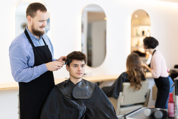 Obraz premium During visit to barbershop, male client communicate with guy stylist, ask to perform haircut, shortening of hair. Hairdresser and visitor discuss hairstyle, master explains and assumes final result