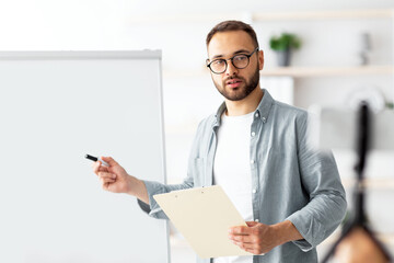 Young male teacher pointing at blackboard with English grammar rules, conducting online lesson from...