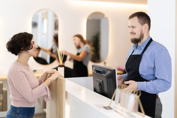 Male stylist stands near barbershop reception desk, communicates with woman visitor, offer various...