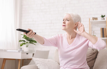 Senior woman cupping her hand behind ear to hear better to the television, making sound louder,...