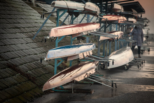 Multiple rowing shells, including sweep oar and coxed four boats, rest on racks by the riverside at sunset. These competitive vessels are used by crews of four or eight. - Powered by Adobe