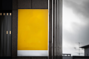 Selective blur on a A bright yellow sign hangs empty outside a modern storefront, awaiting custom branding. The bold, blank panel contrasts with the building’s neutral facade in the city center.