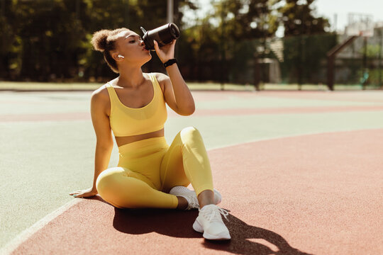 Energy, Vitality, Wellbeing Concept. Fit African American woman in wireless earbuds sitting on basketball field ground, holding shaker, drinking water or protein, taking break after outdoors exercise - Powered by Adobe