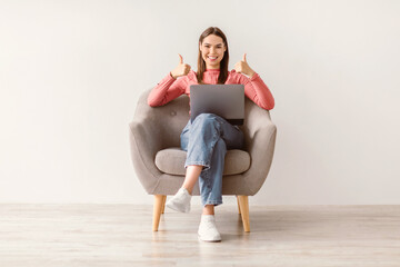 Fototapeta premium Positive young Caucasian woman with laptop working online, sitting in armchair and showing thumbs up with both hands against white studio wall, full length. Remote work and freelancing concept