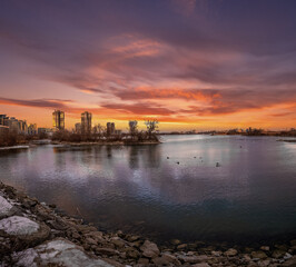 Fototapeta premium Fiery sunset over a calm lake, city skyline in the distance.
