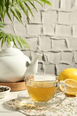 Refreshing green tea in cup, lemon and dry leaves on white table against grey textured wall