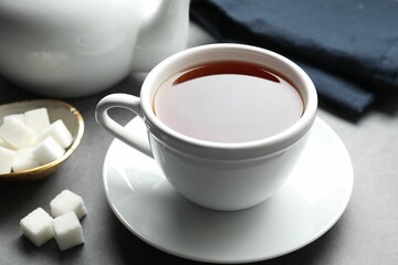 Refreshing black tea in cup and sugar cubes on grey textured table, closeup
