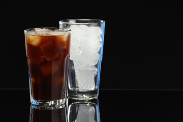 Refreshing iced coffee and ice cubes in glasses on black mirror surface. Space for text