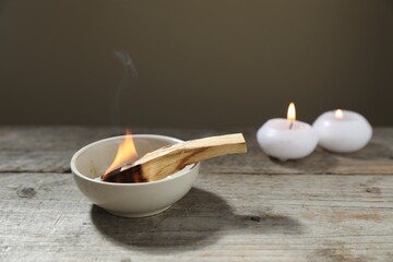 Burning palo santo stick and candles on wooden table against grey background, closeup