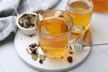 Delicious herbal tea with honey and dry leaves on white tiled table, closeup
