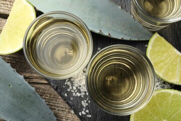 Mexican tequila shots, slices of lime, salt and agave leaves on wooden table, flat lay