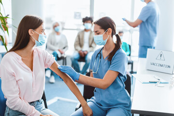 flu Vaccination During Global Pandemic. Young woman in surgical face mask getting injection at hospital. Doctor preparing patient for a shot, rubbing arm with cotton pad, clinic interior