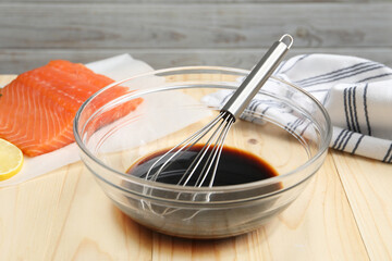 Soy sauce in bowl, whisk and salmon fillet on wooden table, closeup