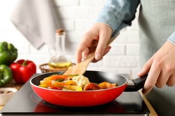 Woman frying vegetables in pan on stove at table indoors, closeup