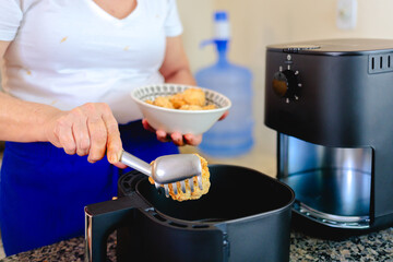 Woman using an air fryer on the countertop of a Brazilian home kitchen. She is placing breaded chicken drumettes into the appliance’s basket to fry them