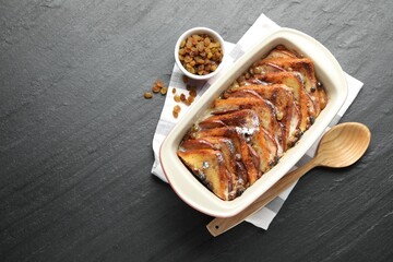 Freshly baked bread pudding in baking dish, raisins and wooden spoon on dark textured table, top view. Space for text
