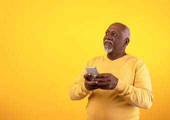 Cheerful senior African American man using mobile phone with new application, looking at empty space on orange studio background. Gadgets and mobile communication concept