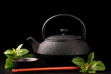 Black cast iron teapot with mint leaves and chopsticks on a black background