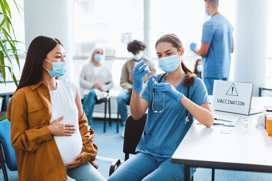 Young woman doctor or nurse in medical face mask and uniform drawing medication out of vial preparing syringe before injecting pregnant asia lady with flu vaccine during vaccination campaign