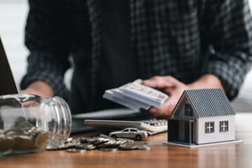 Man counts cash with coins, a calculator, and a miniature house, symbolizing financial planning, savings, and investment management. Savings Plan, Down Payment, Mortgage, Home Equity, Escrow Account.