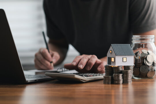 Man calculates finances with a calculator, coins, and a miniature house, symbolizing budgeting, investment, and financial planning. Savings Plan, Down Payment, Mortgage, Home Equity, Escrow Account.