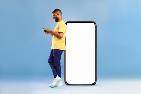Full length of young African American guy using cellphone while leaning on giant mobile phone with empty white screen, blue studio background. Mockup for app or website, space for advertisement
