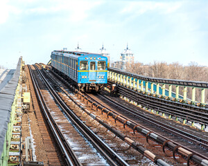 Fototapeta premium A vibrant blue Kyiv metro train traveling across a bridge, framed by snowy tracks and city architecture in the distance.