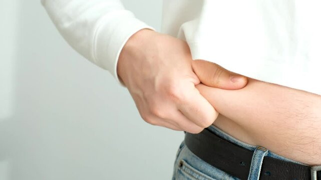 A male hand holding excessive belly fat on gray background, overweight concept.