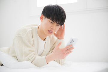 スマホを見てうつ伏せで寝転ぶ困る、迷う、悩む表情の若い20代の男性
