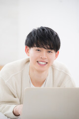 ベッドの上でうつ伏せで寝転がりパソコンをする笑顔の20代の若い男性カメラ目線　縦　学生やフリーランスの若手のイメージ