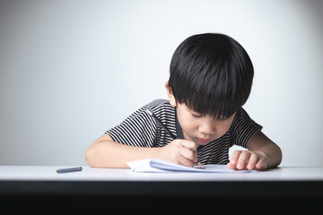 A focused Thai boy writing in a notebook with a pencil, concentrating on his studies at a desk.