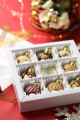 Box with tasty chocolate candies in shape of baubles and Christmas decor on red background, closeup
