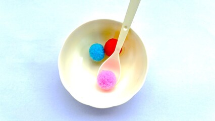 Bright red, blue, and pink soft pom-pom balls placed on white plate arranged on a white surface. Ideal for craft, DIY, art projects, kids’ toys, and creative design concepts in stock photography.