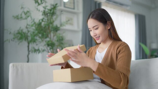 Happy young Asian woman opening delivery package at home. Beautiful female customer open box and sitting on couch in living room