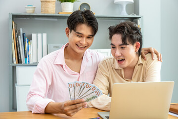 Obraz premium Two Asian men gay couple celebrating financial success with cash while working on laptop at home