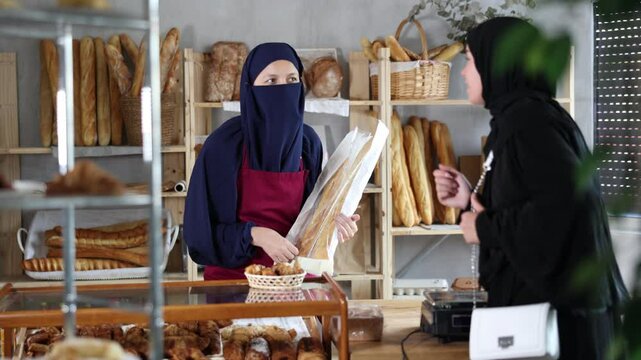 Woman in niqab and apron working at counter in cozy bakery filled with various breads and pastries, handing bag with freshly baked baguettes to female customer in black hijab. High quality 4k footage