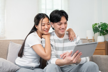 A lovely Asian couple sits together on a sofa in the living room, watching a video on a tablet.