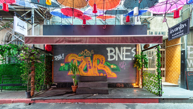 Istanbul, Turkey - May 13, 2023: Group of colorful umbrellas hanging over a closed shop rolling door with colorful graffiti in a narrow alley in Karakoy neighborhood
